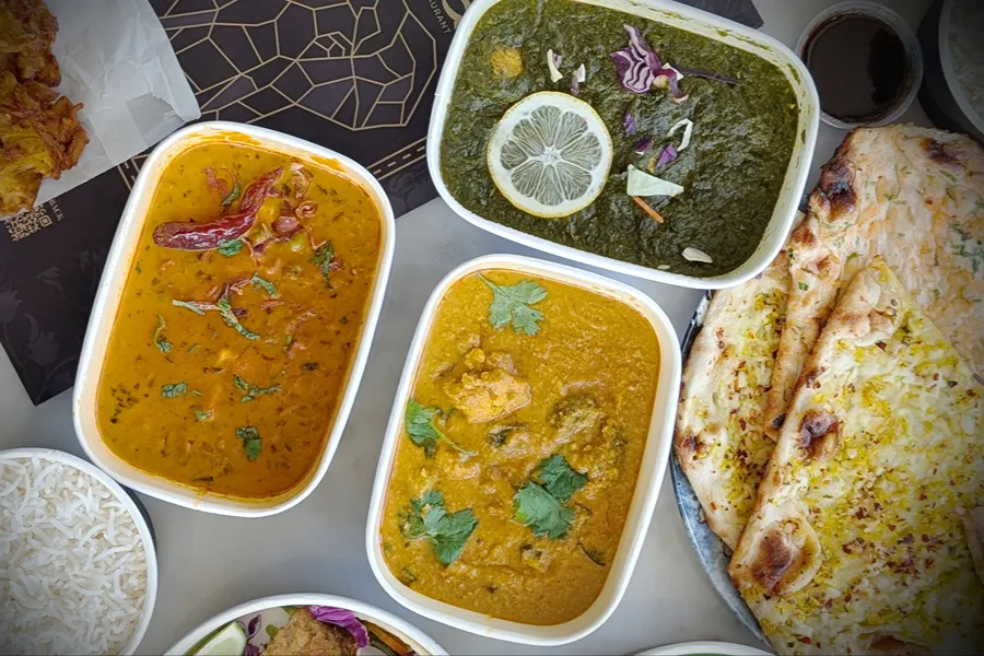 Saag and two curries packed in Sula takeout containers beside fresh garlic naan and a bowl of basmati rice.