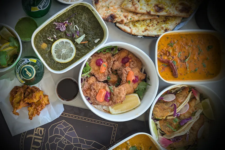 An Indian takeout spread from Sula in Vancouver, with saag, curries, garlic naan, basmati rice and pakoras laid out in takeaway containers.
